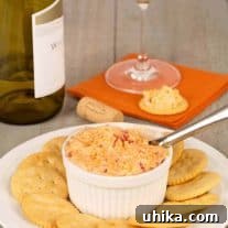 A white crock filled with pimento cheese on a plate surrounded by crackers, a wine bottle, wine glass, and cracker topped with cheese in the background.