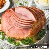 A large, spiral-sliced, glazed ham on a serving platter, surrounded by parsley garnish, mashed potatoes and a striped tea towel in the background, ready for a festive meal.