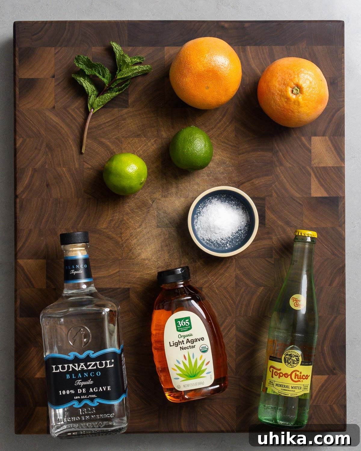 The Minted Classic Paloma 3 A flat lay photograph showcasing all the fresh and essential ingredients for making a Paloma cocktail: vibrant mint leaves, ripe grapefruits, fresh limes, a pinch of salt, a bottle of blanco tequila, agave nectar, and sparkling water.