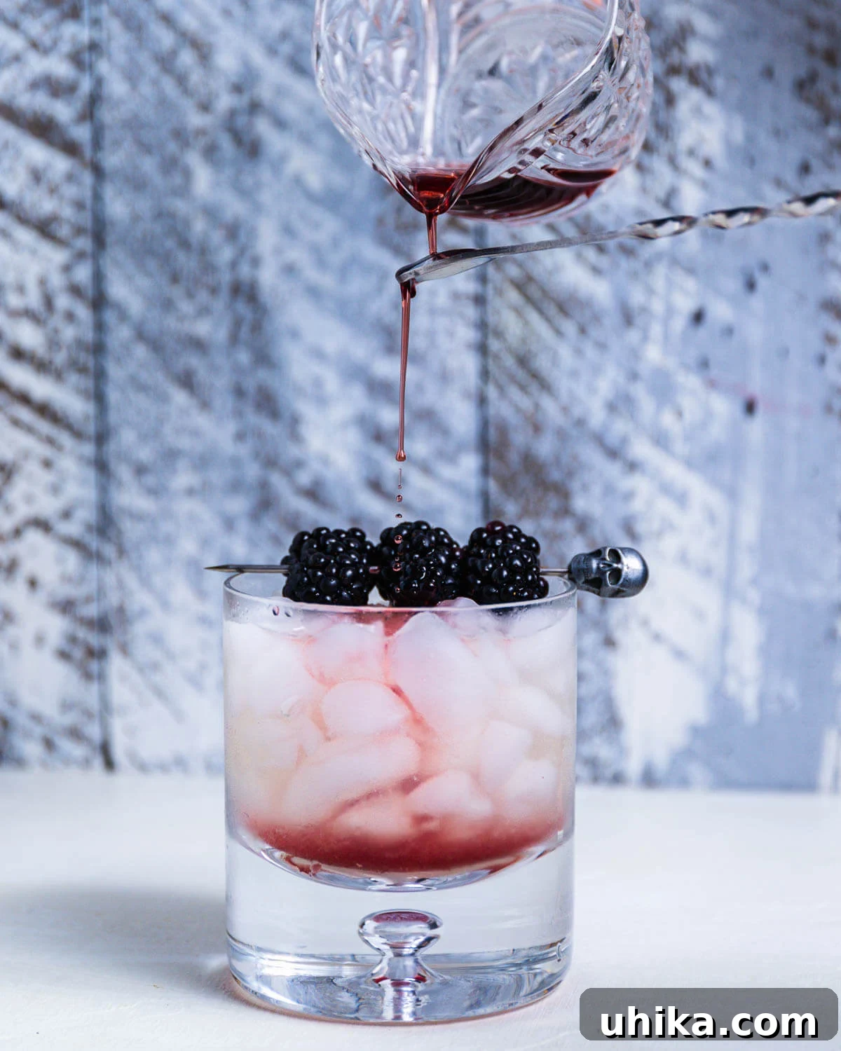 Blackberry Bramble - sipandfeast.com #bramble #drinks #cocktails A bartender's hand expertly drizzling crème de mûre over the back of a bar spoon into a rocks glass filled with crushed ice, creating a marbling effect.
