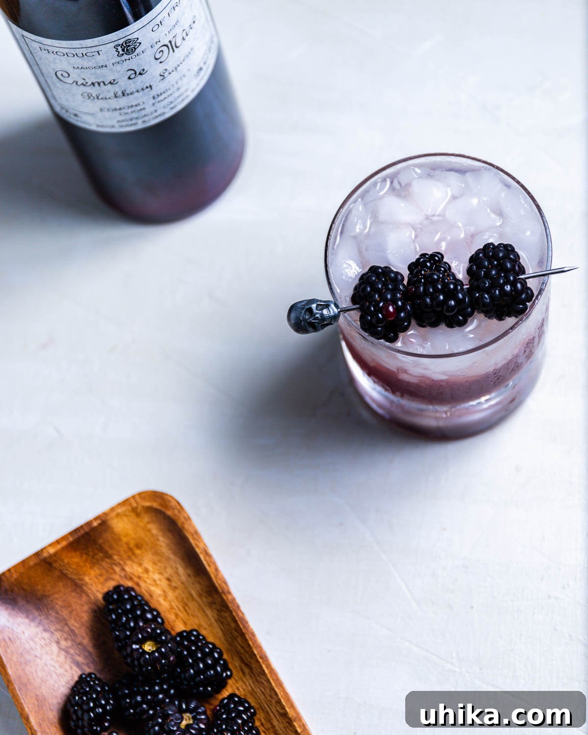 Blackberry Bramble - sipandfeast.com #bramble #drinks #cocktails An overhead shot of a finished Blackberry Bramble cocktail, adorned with fresh blackberries on a skull-themed skewer, with a bottle of crème de mûre and a plate of loose blackberries in the background.