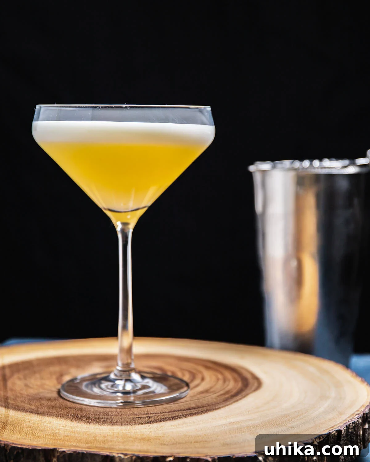Oliveto cocktail in coupe glass on round wooden board with metal shaker in background, showcasing its frothy top.