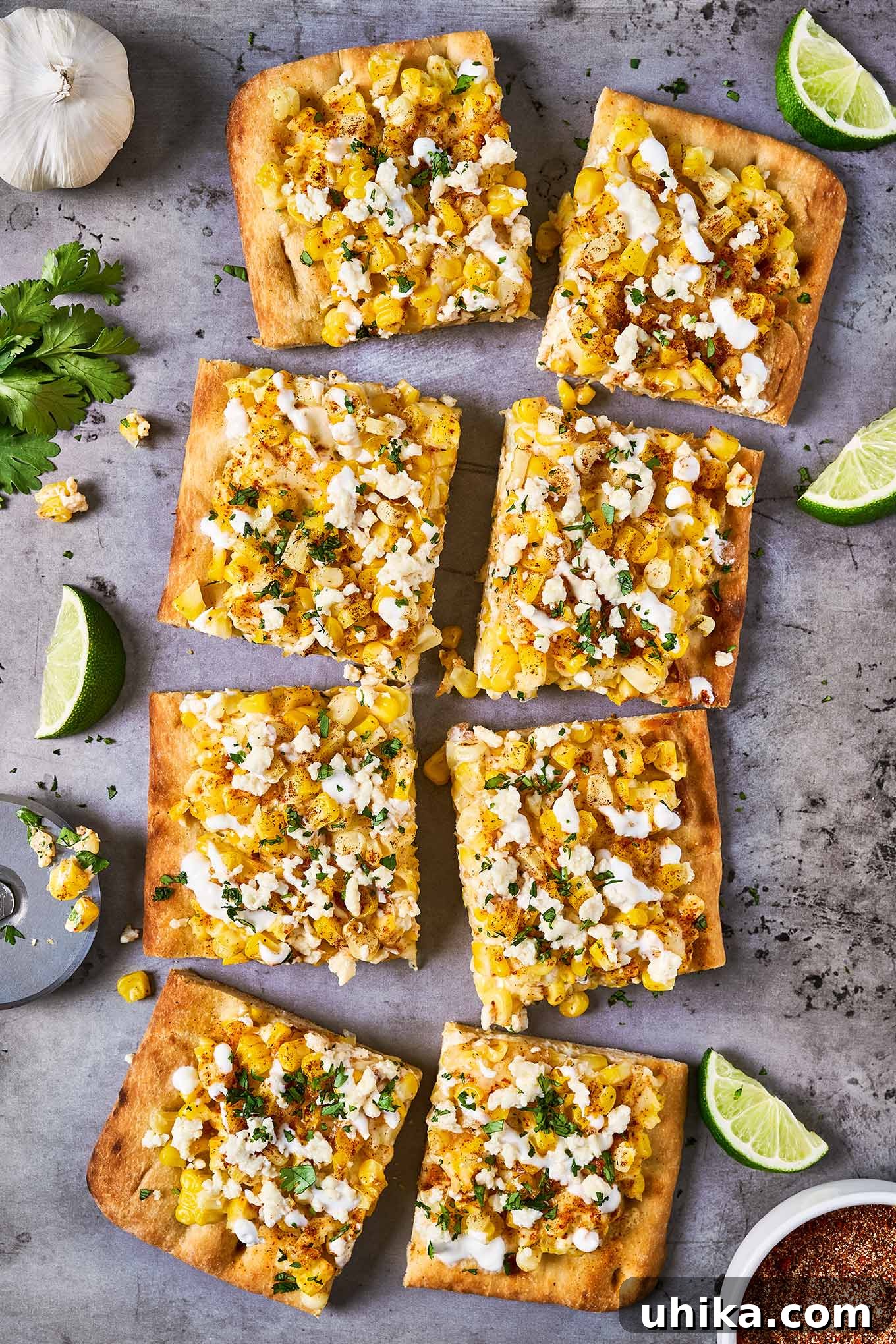 Elote Flatbread Fiesta 2 Vibrantly seasoned Mexican street corn flatbread, perfectly cut into squares, garnished with fresh cilantro, and served on a baking sheet alongside zesty lime wedges, minced garlic, and a sprinkle of elote seasoning.