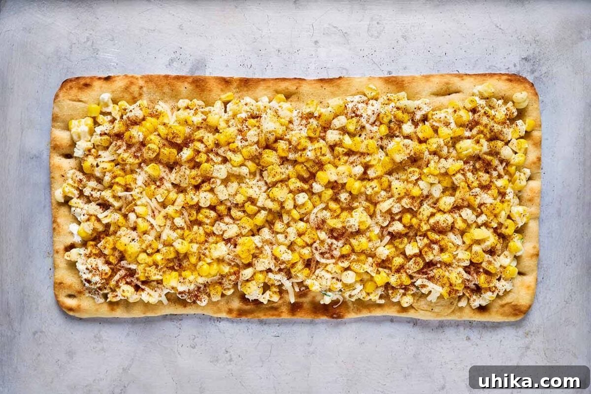 Elote Flatbread Fiesta 5 A rectangular flatbread, generously topped with a vibrant mixture of fresh corn, mayonnaise, sour cream, crumbled queso fresco, shredded mozzarella, and a sprinkle of authentic elote seasoning, awaiting its transformation in the oven on a baking sheet.