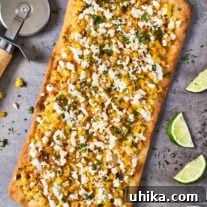 Elote Flatbread Fiesta 7 A flatbread topped with mozzarella cheese, corn kernels, queso fresco, chopped cilantro, seasoning blend, and crema on a baking sheet.