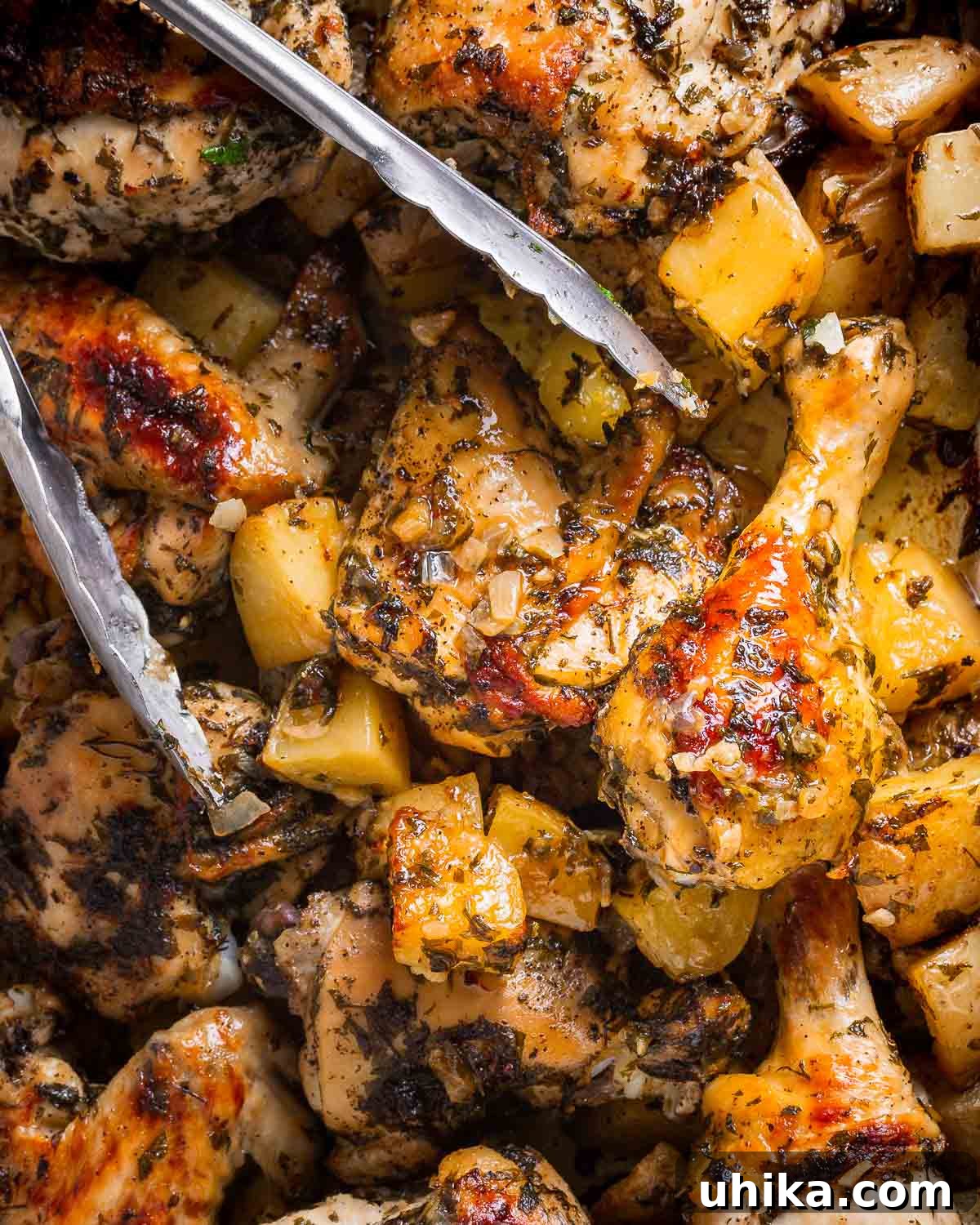 Garlic Rosemary Chicken and Roasted Potatoes 2 Tongs gently lifting a piece of perfectly baked Italian chicken from a pan filled with roasted potatoes.