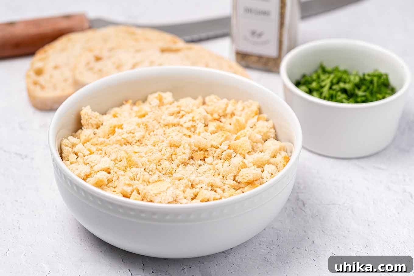 Fresh, homemade breadcrumbs in a bowl with a slice of bread, a prep bowl of chopped parsley, a bread knife, and some seasoning in the background.
