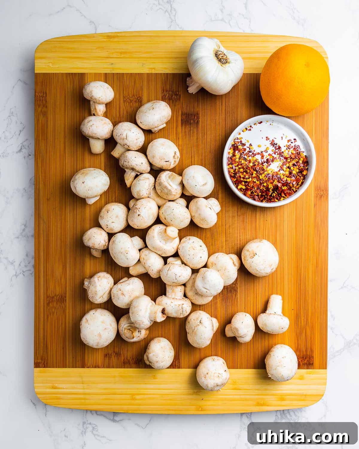 Flavor Infused Mushrooms 3 Ingredients for marinated mushrooms: white button mushrooms, whole garlic cloves, hot red pepper flakes, and a whole orange on a rustic wood cutting board.