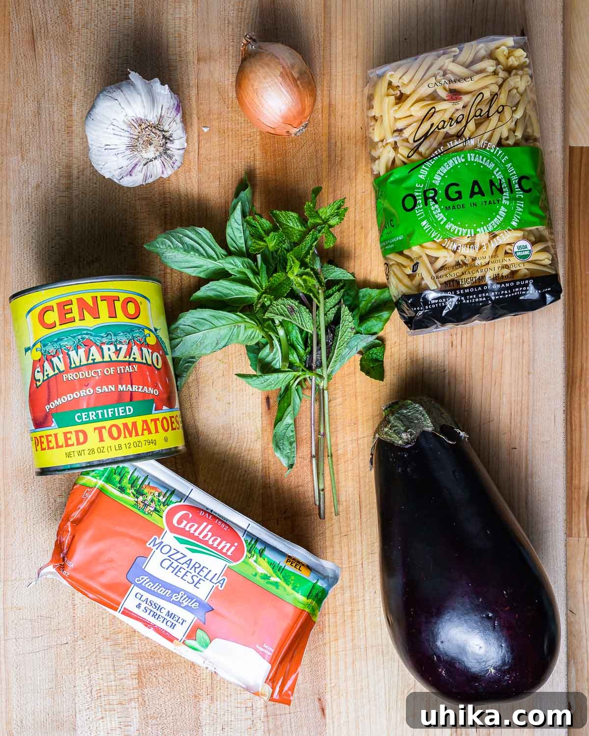 Ingredients for Pasta alla Siciliana displayed on a countertop: garlic cloves, a whole onion, casarecce pasta, a can of plum tomatoes, fresh basil, fresh mint, a block of mozzarella, and a large eggplant.