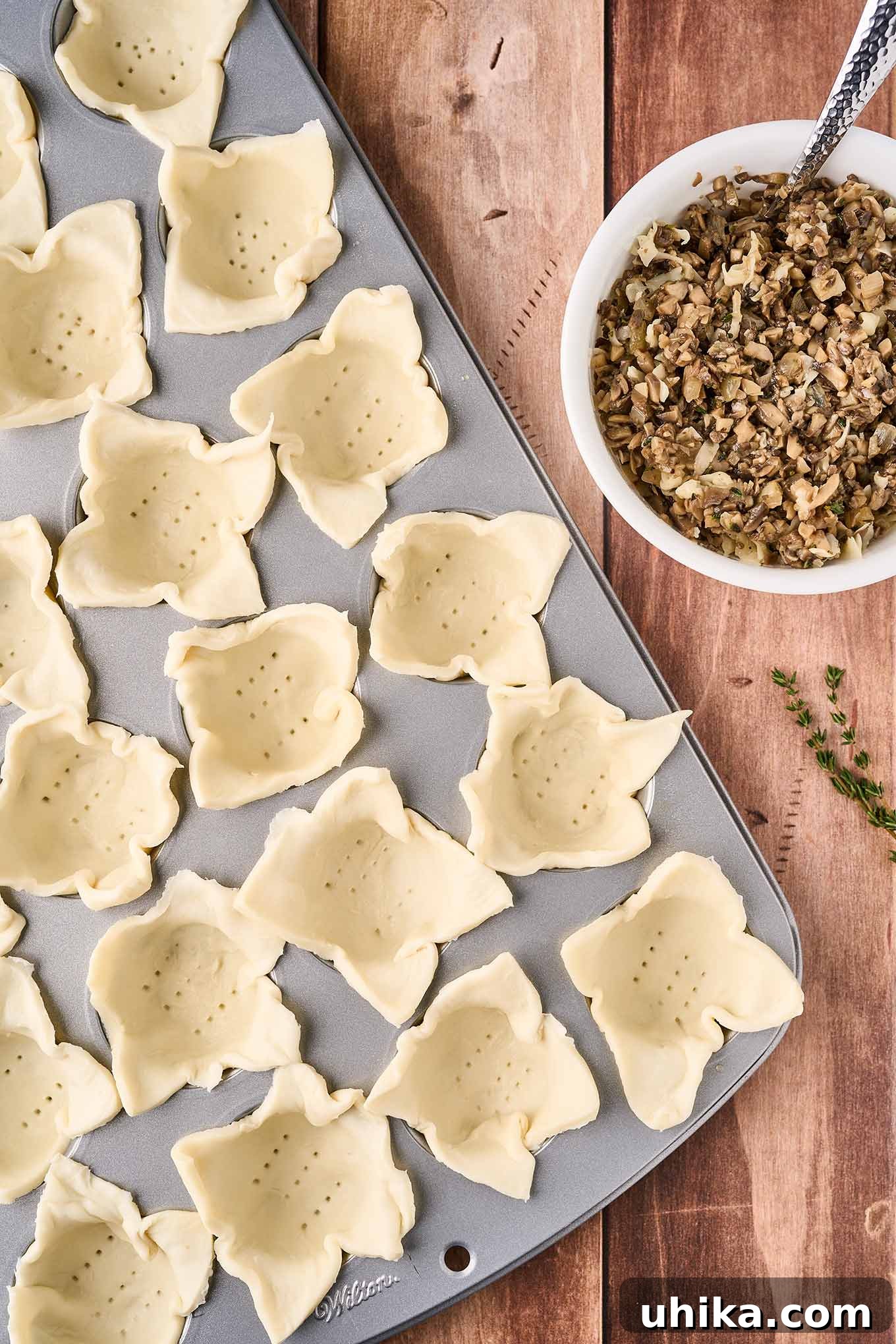 Umami Cloudlets 6 A mini muffin tin filled with squares of puff pastry, perforated on the bottom and ready to be filled with the sautéed mushroom mixture in a bowl on the right.