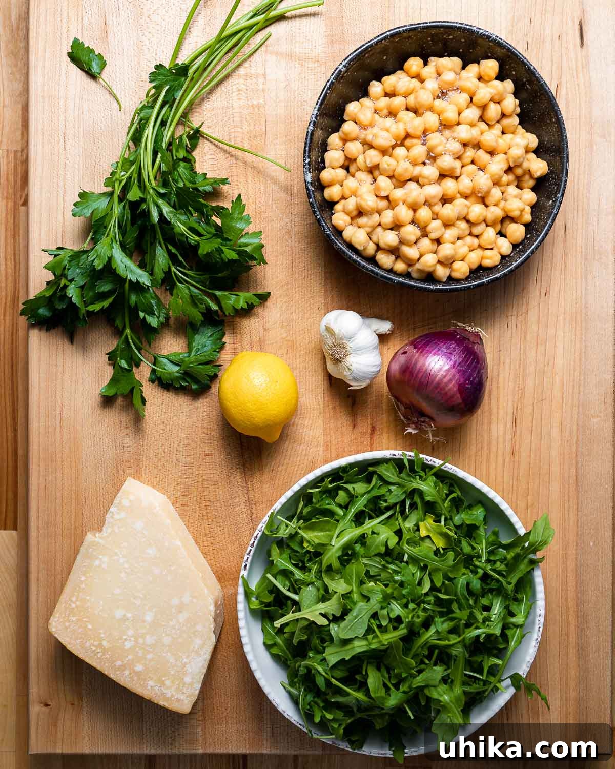 Zesty Lemon Parmesan Chickpea Arugula Salad 3 Key ingredients for the chickpea arugula salad including fresh parsley, chickpeas, lemon, garlic, red onion, a block of Parmesan cheese, and a bowl of arugula.