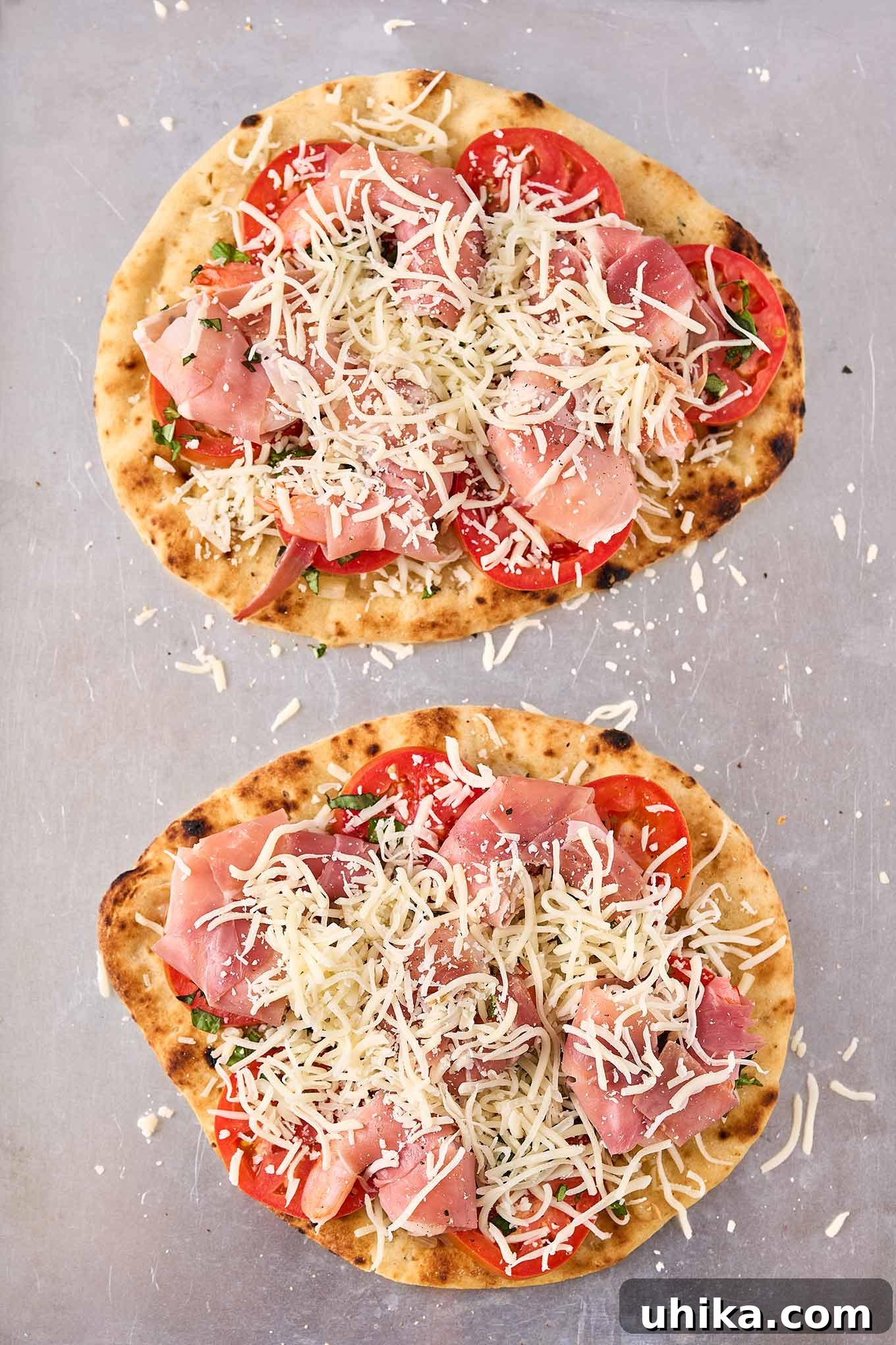 Prosciutto-Wrapped Shrimp Naan Pizza 4 Two naan flatbread pizzas on a baking sheet, topped with prosciutto-wrapped shrimp, tomatoes, and mozzarella and ready for the oven.