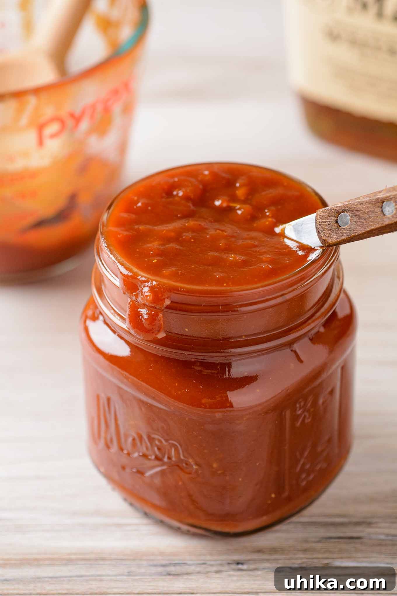 Handcrafted Bourbon BBQ Delight 2 A small jar of rich, homemade bourbon barbecue sauce with a wooden-handled spoon, measuring cup, and a bottle of bourbon in the background.