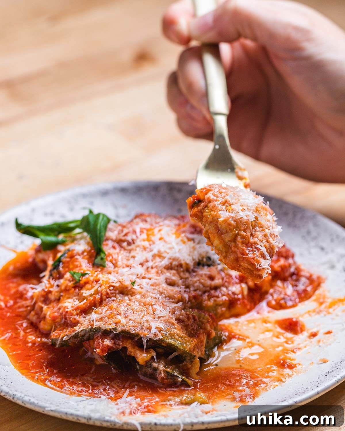 Fork lifting a piece of Zucchini alla Parmigiana from a baking dish, with a serving on a white plate below.