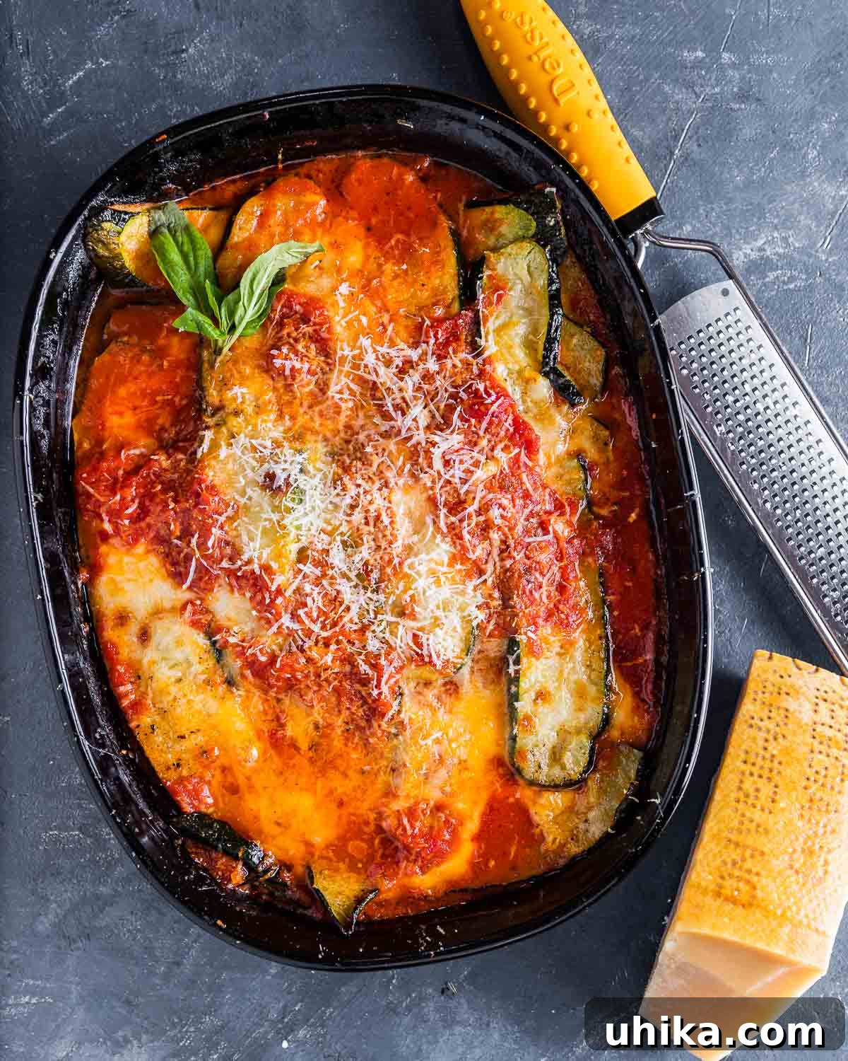Overhead shot of freshly baked Zucchini alla Parmigiana in a baking dish, with a block of Parmigiano Reggiano cheese and fresh basil leaves on the side.