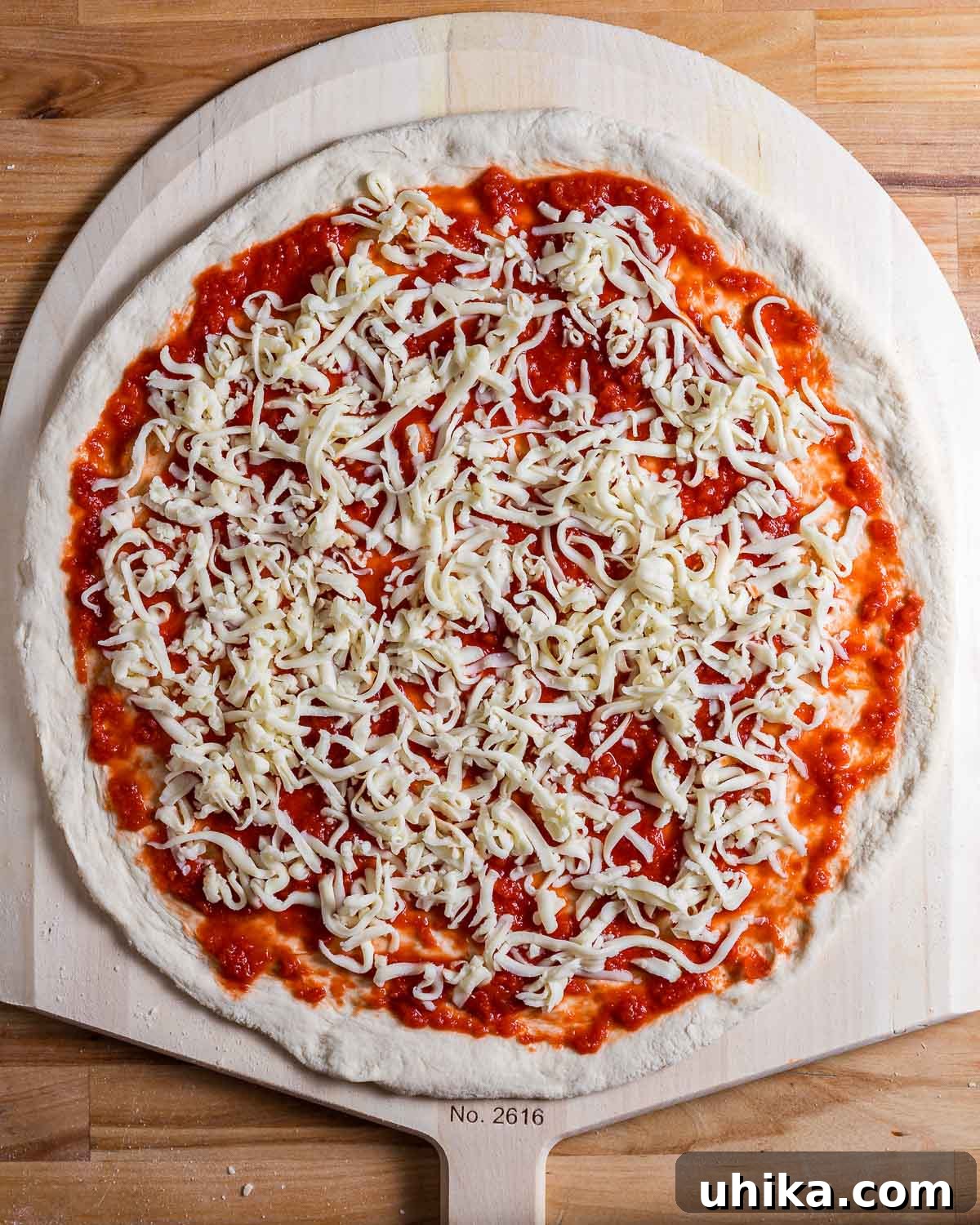 DIY Pizza Steel: Crafting Your Own Perfect Crusts 7 Uncooked pizza dough topped with sauce and cheese resting on a wooden pizza peel, ready for baking.