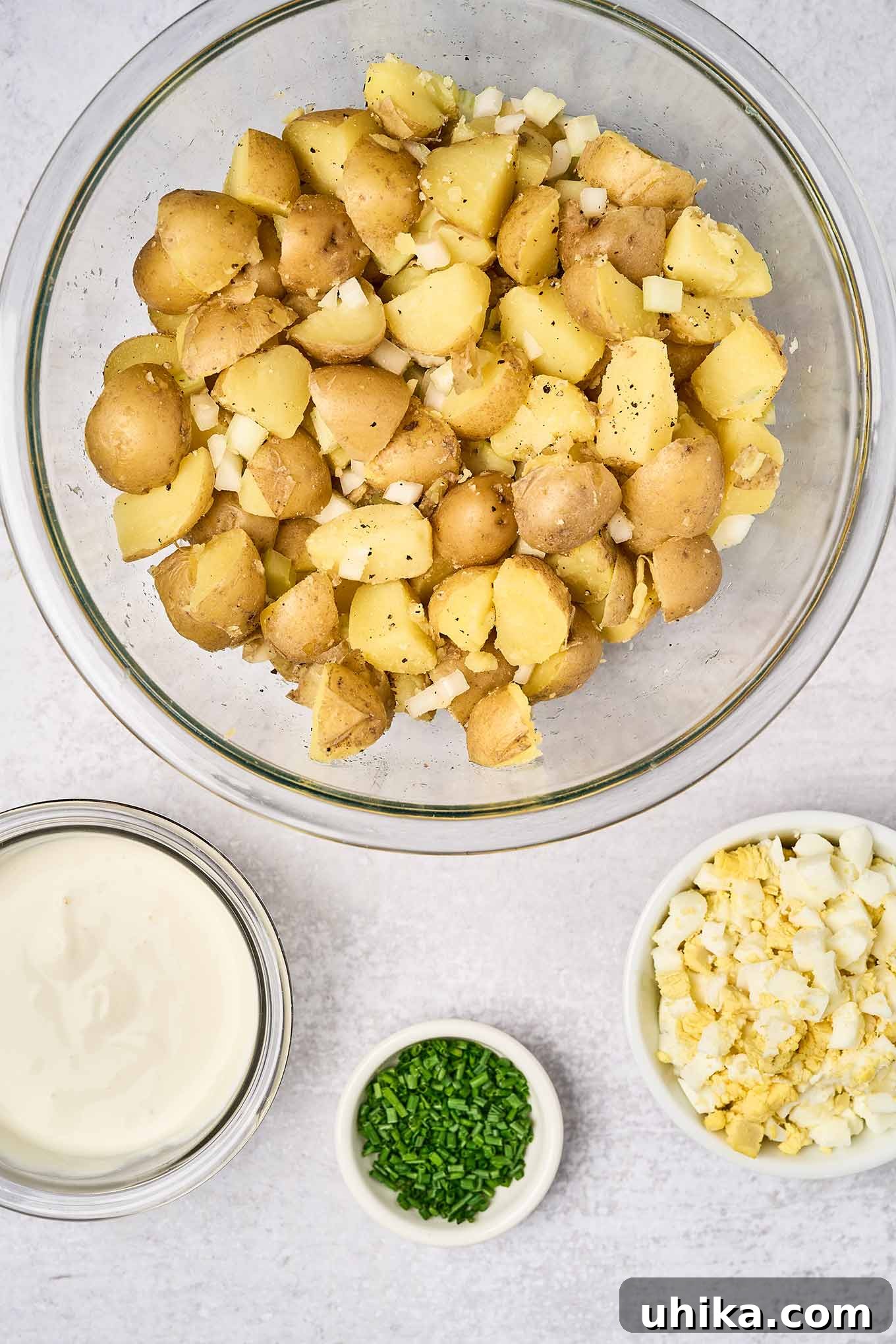 Zesty Creamy Potato Salad 4 Boiled chunks of baby yellow potatoes and chopped celery and onion in a mixing bowl next to a small bowl of chopped hard boiled eggs, a prep bowl of chives, and a bowl of sour cream dressing - all ready to be combined.