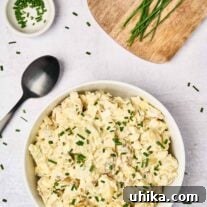 Sour cream potato salad in a white serving bowl, garnished with fresh chives, a black serving spoon to the left, a prep bowl of chopped chives, and fresh chives on a wooden board in the background.