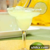A classic margarita on the rocks in a clear margarita glass, garnished with a lime wedge. A whole lime is on the table in the foreground, remaining margaritas in a pitcher in the background.