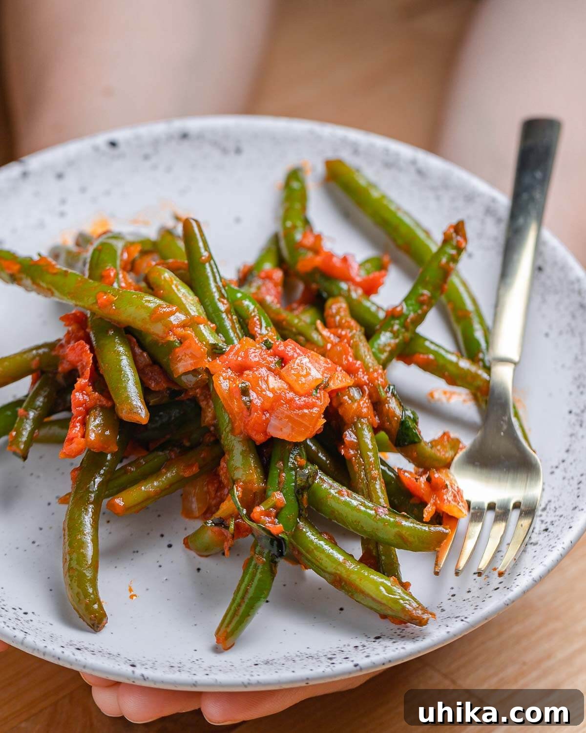 Tender Green Beans Simmered in Rich Tomato Sauce 8 A white plate held in hands, showcasing a serving of perfectly stewed green beans with tomato sauce, garnished with fresh herbs and ready to be enjoyed.