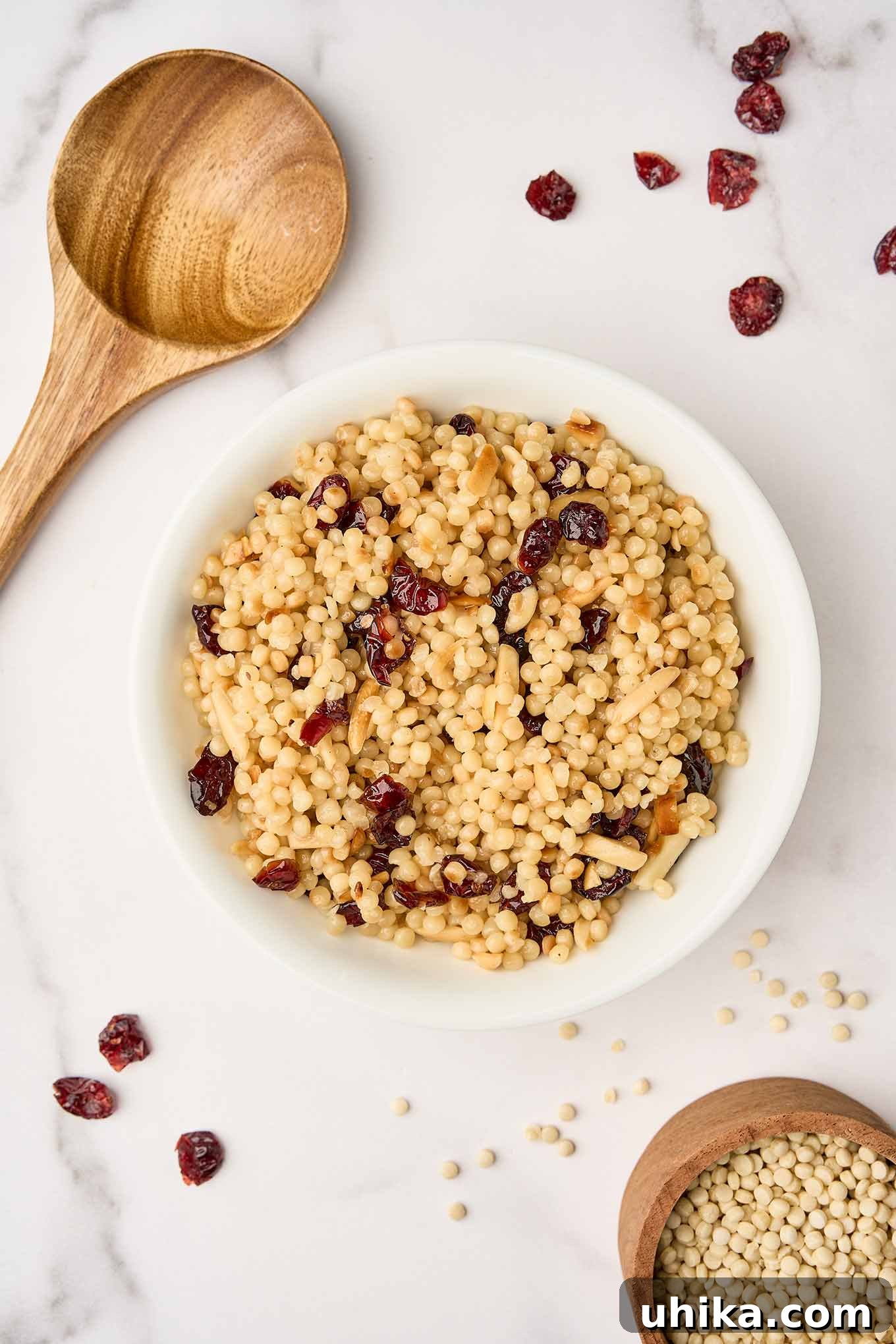 Sweet and Tart Pearl Couscous with Almond Crunch 2 A vibrant bowl of toasted pearl couscous, generously mixed with ruby-red dried cranberries and golden slivered almonds, sits alongside a wooden spoon, scattered cranberries, and a measuring cup filled with uncooked Israeli couscous.