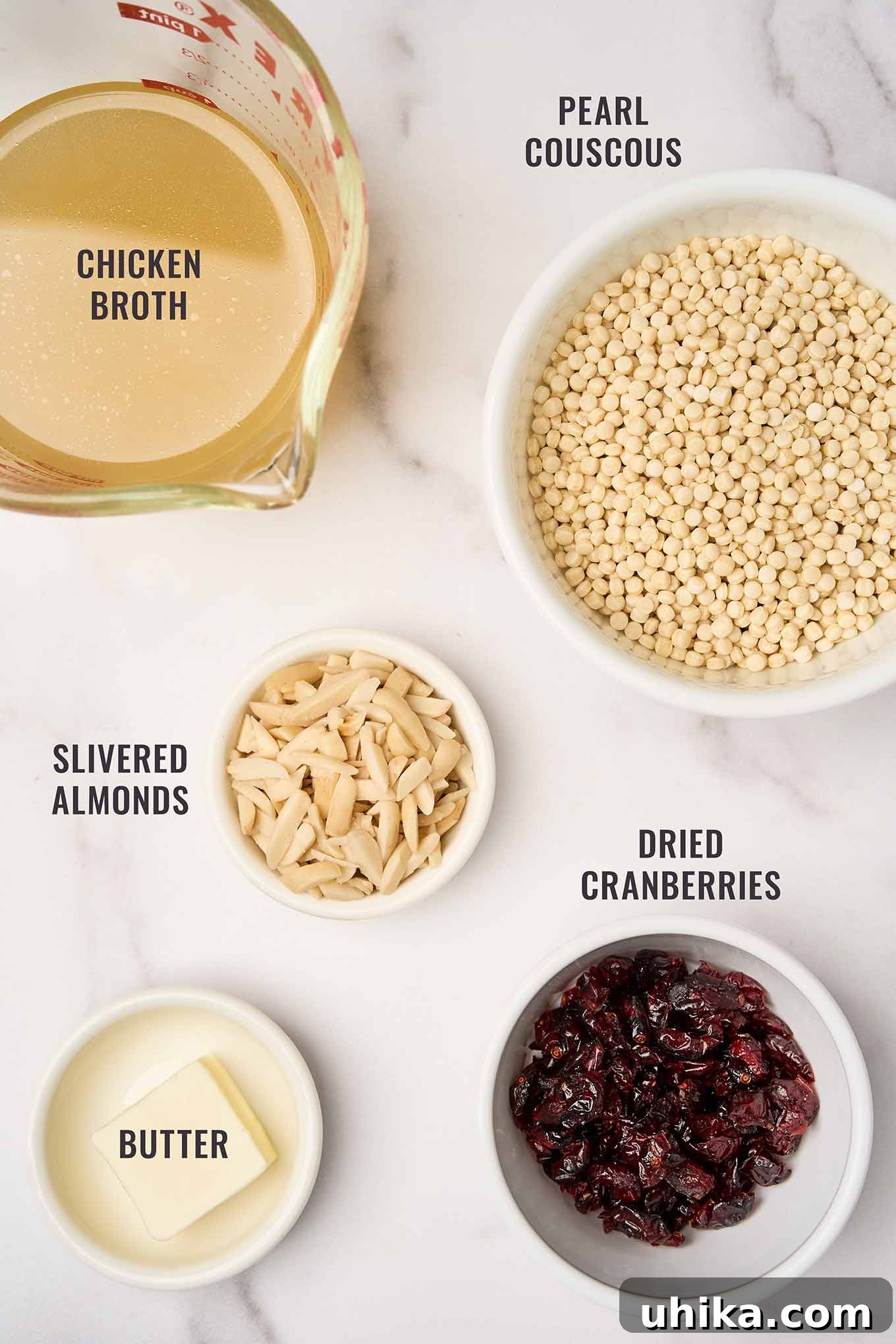 Sweet and Tart Pearl Couscous with Almond Crunch 3 An organized, overhead view of the key ingredients required to prepare a delicious pearl couscous dish with cranberries and almonds, showcasing pearl couscous, chicken broth, butter, slivered almonds, and dried cranberries.
