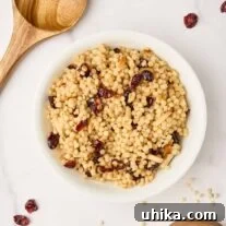 A bowl of toasted pearl couscous with dried cranberries and slivered almonds surrounded by a wooden spoon, scattered cranberries, and a wooden measuring cup with uncooked Israeli couscous.