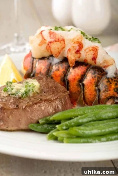 A closeup view of a pan-seared filet mignon topped with parsley butter next to a lobster tail with its juicy meat on top, and a serving of green beans.