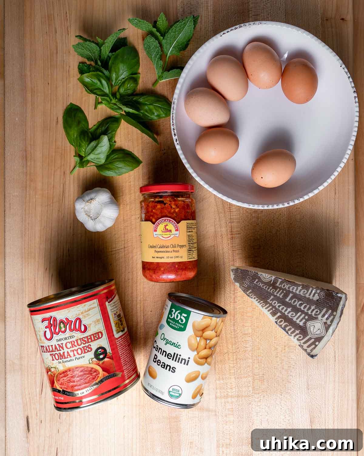 A vibrant flat lay image showcasing all the fresh ingredients for Eggs in Purgatory: eggs, fresh basil, mint, garlic cloves, a jar of Calabrian chili paste, canned tomatoes, cannellini beans, and grated Pecorino Romano cheese.