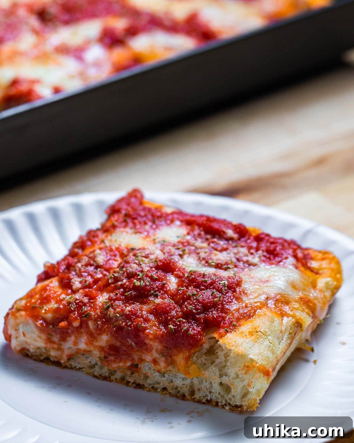 A perfectly triangular slice of New York Sicilian pizza, showcasing its thick crust and generous toppings, on a white plate.