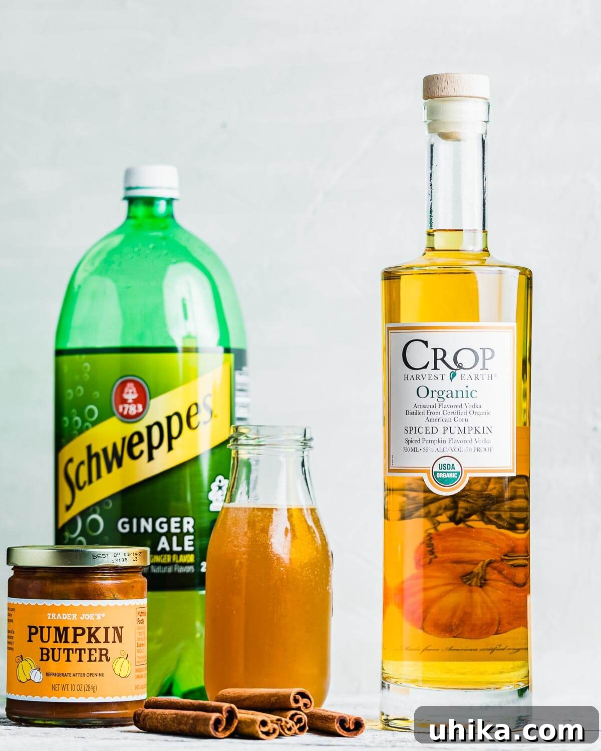 Spiced Pumpkin Harvest Mule 4 All ingredients for Pumpkin Mule shown on a dark surface: a bottle of ginger ale, a bottle of apple cider, cinnamon sticks, and a jar of pumpkin butter.