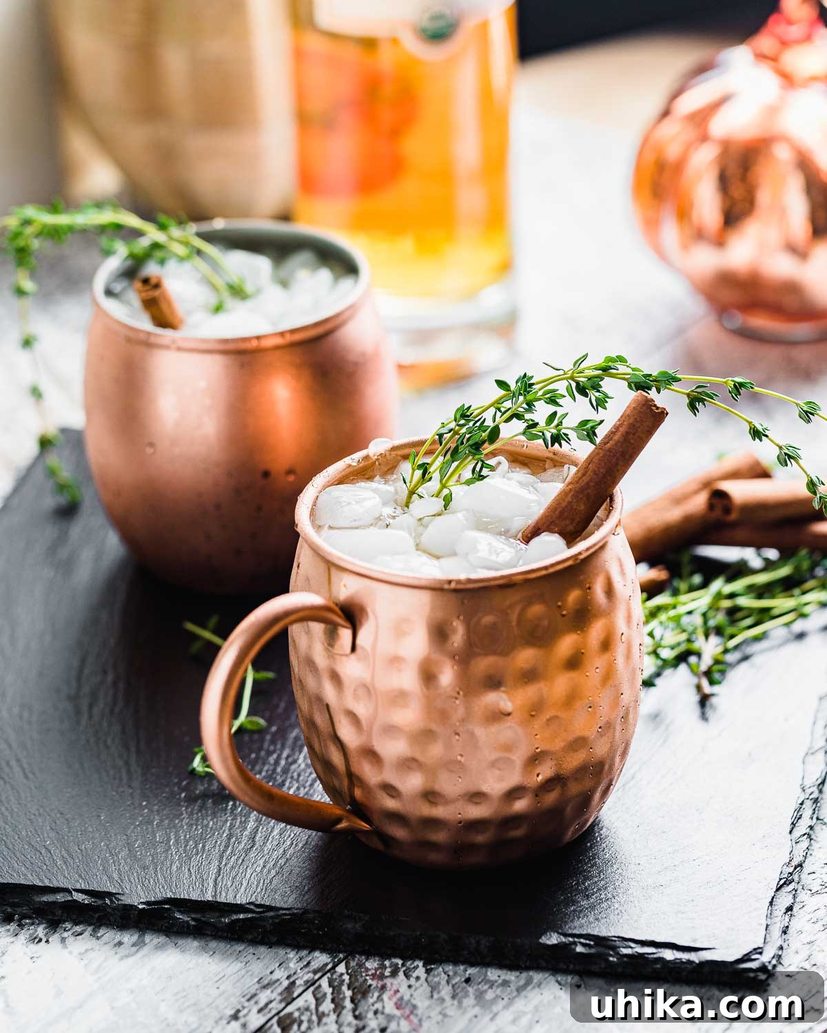 Spiced Pumpkin Harvest Mule 3 A Pumpkin Mule cocktail in a copper mug, garnished with a cinnamon stick and thyme, with a blurred background of autumn decorations.