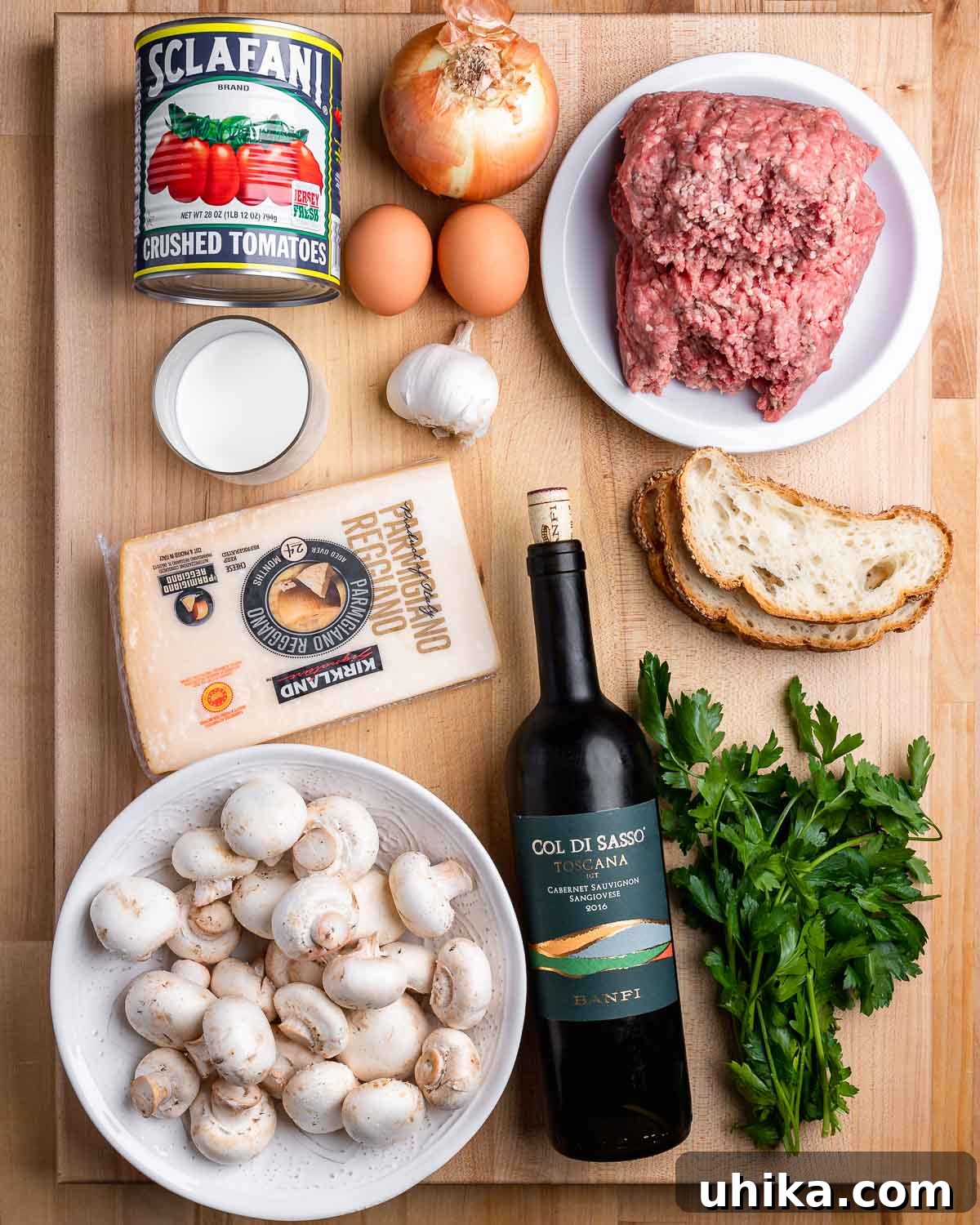 A collage of fresh ingredients for Italian meatloaf: plum tomatoes, onion, garlic cloves, eggs, ground beef, slices of bread, parmesan cheese, mushrooms, red wine, and fresh parsley.