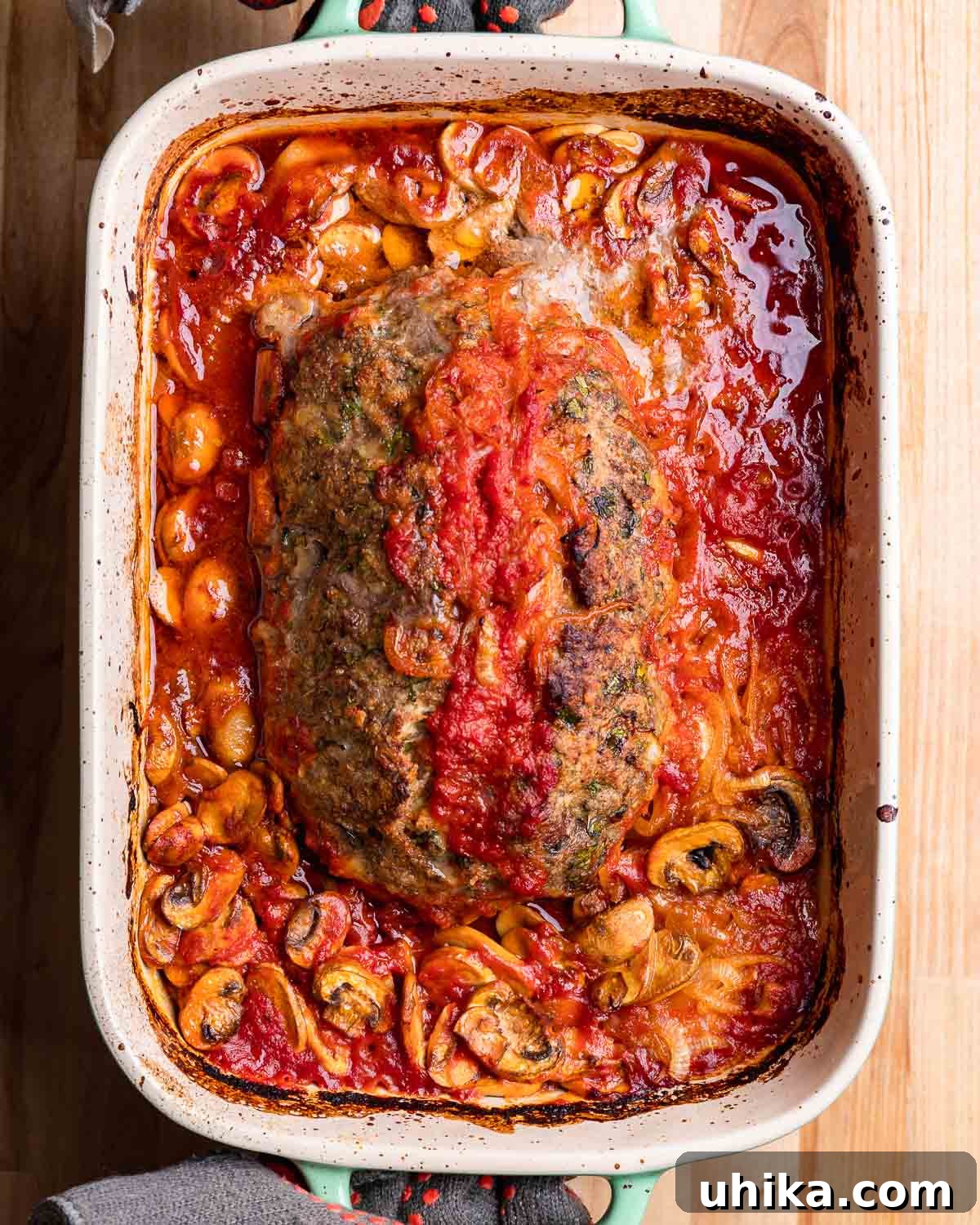 Gloved hands carefully removing a green baking dish containing the freshly cooked Italian meatloaf with mushrooms and sauce from the oven.