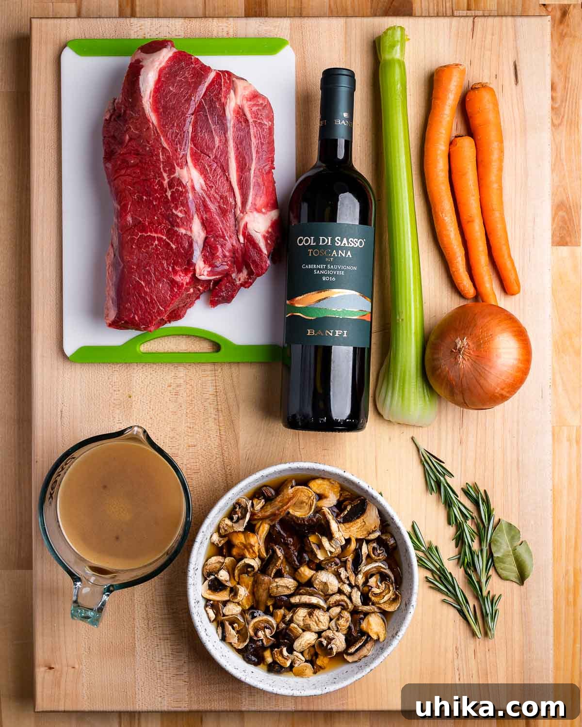 Hearty Italian Beef Stew 3 Ingredients for Italian beef stew laid out: cubed beef chuck, bottle of red wine, chopped celery, carrots, and onion, beef stock, fresh mushrooms, and a sprig of rosemary and bay leaf.