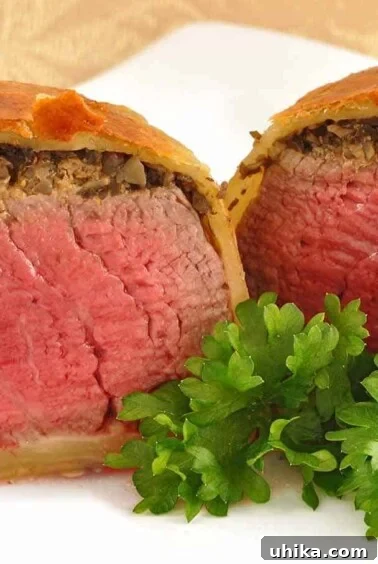 A dinner plate with an individual beef Wellington, sliced in half to show the perfectly cooked beef, topping of mushroom duxelles, and flaky pastry wrap.