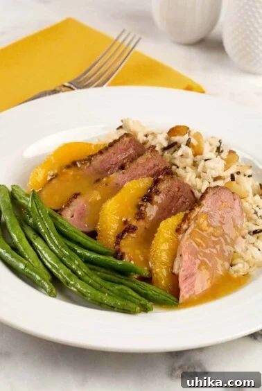 A plate with a serving of duck breast a l'orange; sliced duck breast, medium rare, orange supremes, rice pilaf, and green beans.