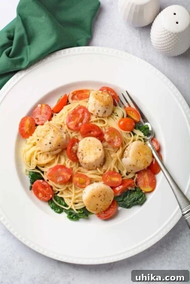 Scallops and pasta with white wine butter sauce, angel hair, tomatoes, and sautéed spinach on a white plate.