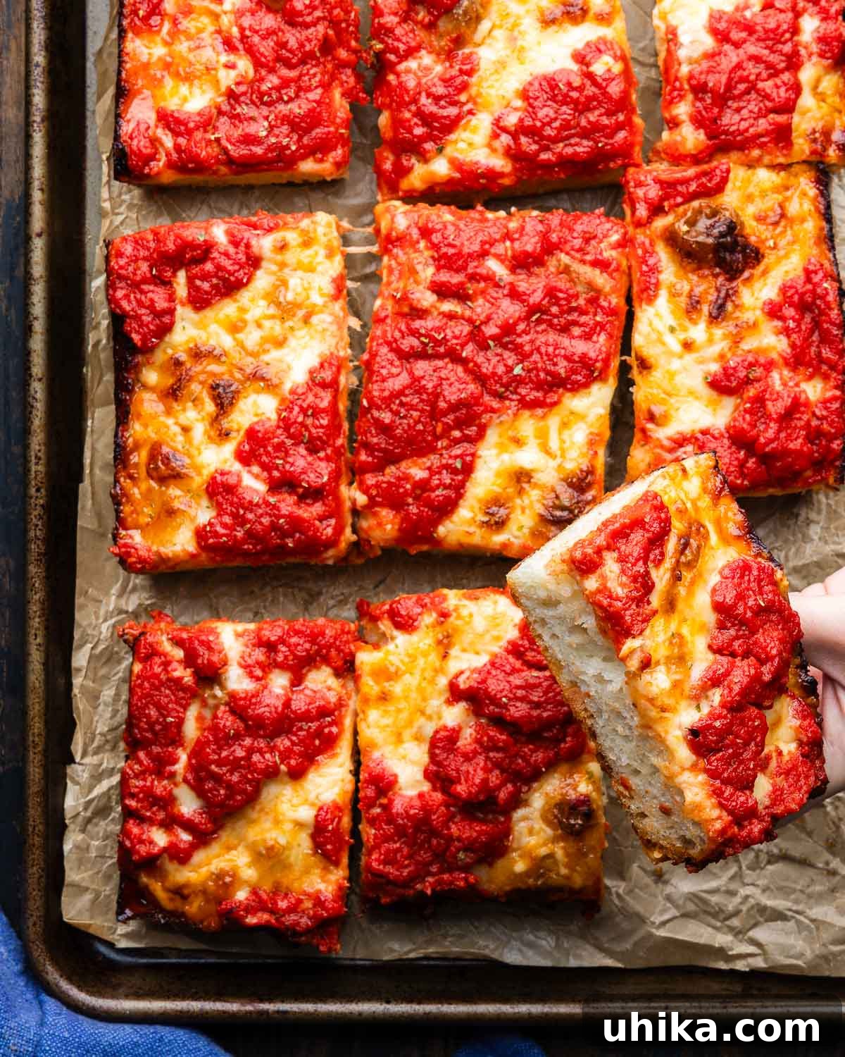 Pillowy Detroit Pizza 2 Detroit pizza in pan with one slice held in hands, showcasing its thick, crispy crust and rich toppings.