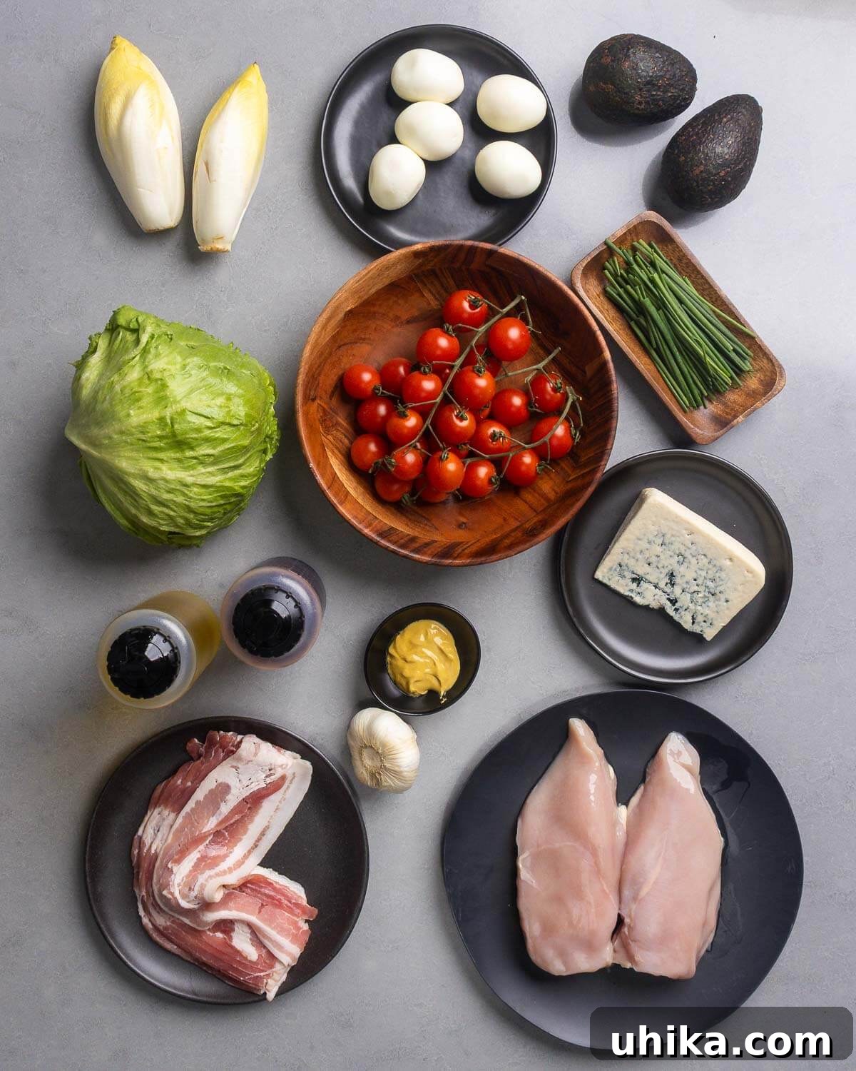 Hearty Cobb Salad Recipe with Avocado and Bacon 4 Ingredients shown: endive, eggs, avocados, lettuce, tomatoes, chives, blue cheese, olive oil, vinegar, dijon mustard, garlic, bacon, and chicken breasts.