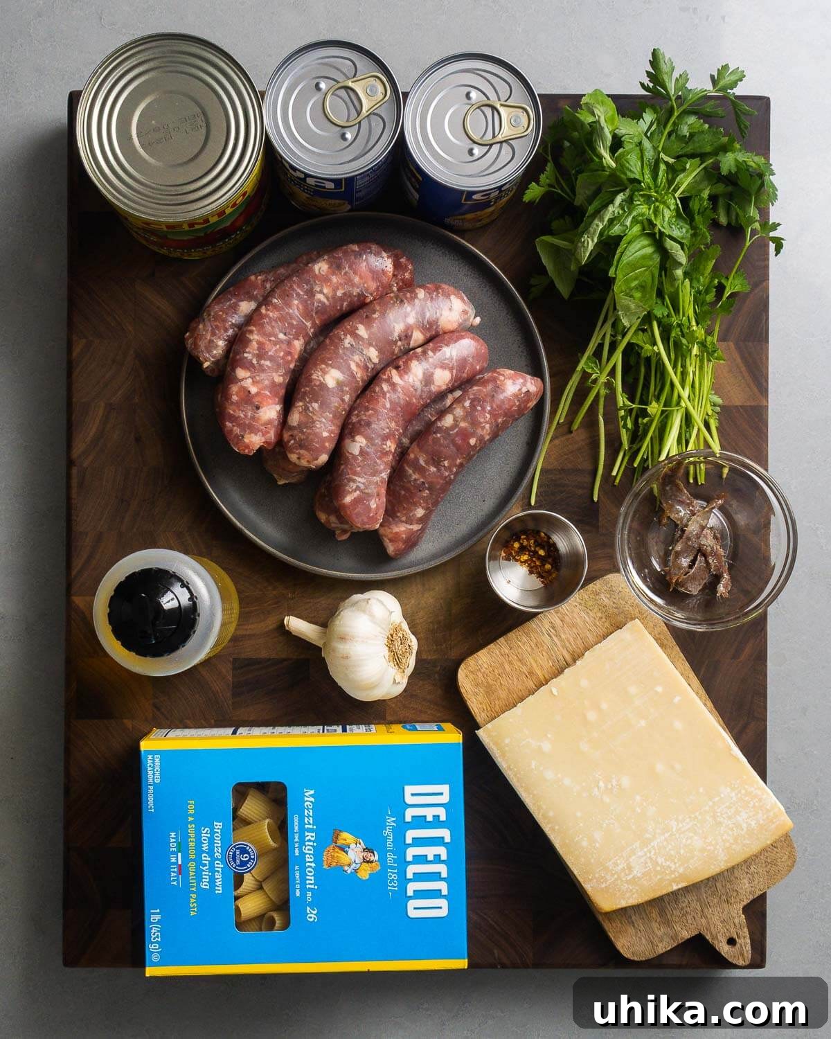 One-Pot Sausage and Chickpea Pasta 4 Ingredients shown: canned tomatoes, canned chickpeas, basil, parsley, Italian sausage, hot red pepper flakes, anchovies, olive oil, garlic, rigatoni pasta, and block of Parmigiano Reggiano.
